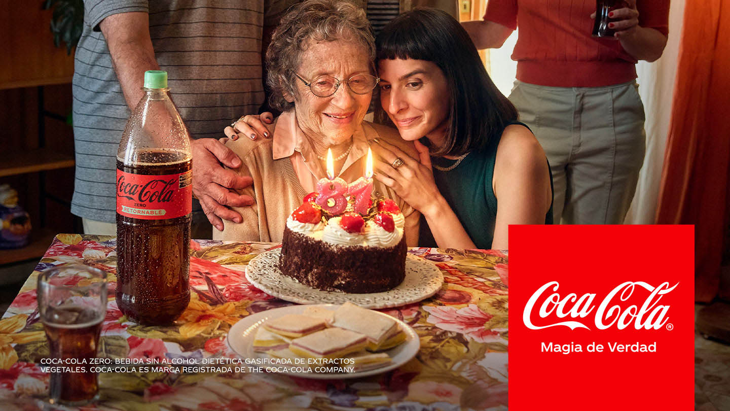 Una familia reunida festeja el cumpleaños de la abuela. Una torta en el centro de la mesa. Una nieta que abraza a su abuela. Hay una Coca-Cola Zero Retornable al costado de la mesa.