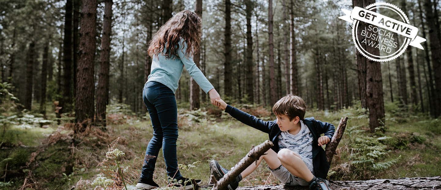 Ein Mädchen reicht einem Jungen, der am Baumstamm im Wald sitzt, die Hand und hilft ihm auf.