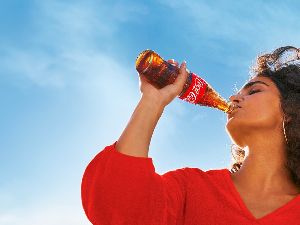 Uma mulher de camisa vermelha aprecia uma Coca-Cola em uma garrafa de vidro retornável sob um céu azul.