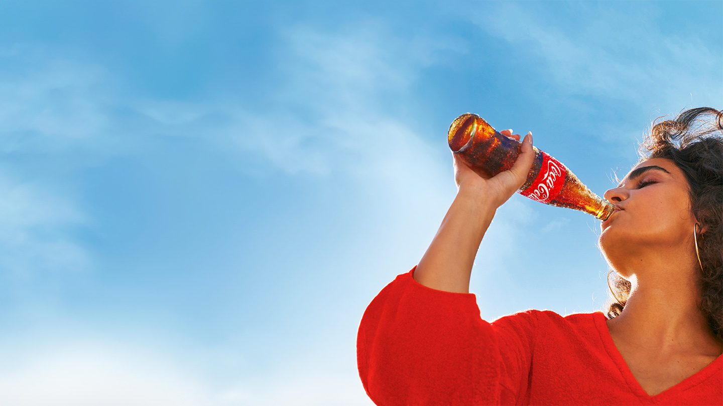 Uma mulher de camisa vermelha aprecia uma Coca-Cola em uma garrafa de vidro retornável sob um céu azul.