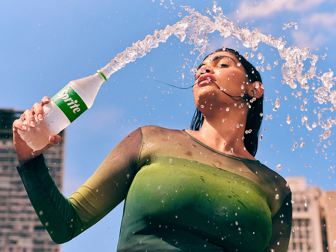 Mulher vestindo camisa verde despeja Sprite gelada de uma garrafa sobre a cabeça, aproveitando a refrescância em um dia quente de verão na cidade.