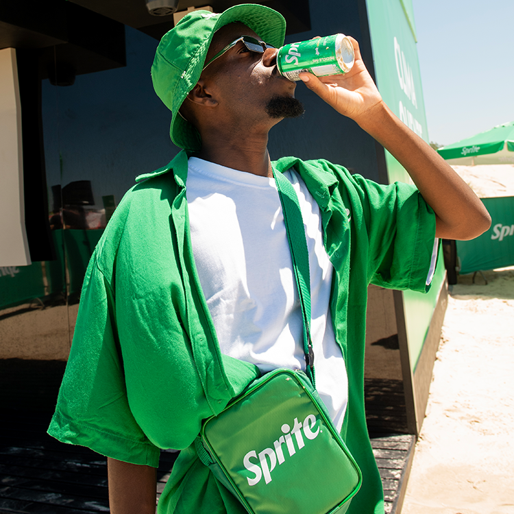 Um homem usando chapéu bucket verde, óculos escuros, camisa verde e camiseta branca bebe Sprite diretamente da lata na praia. Ele também usa uma pequena bolsa verde da marca Sprite.