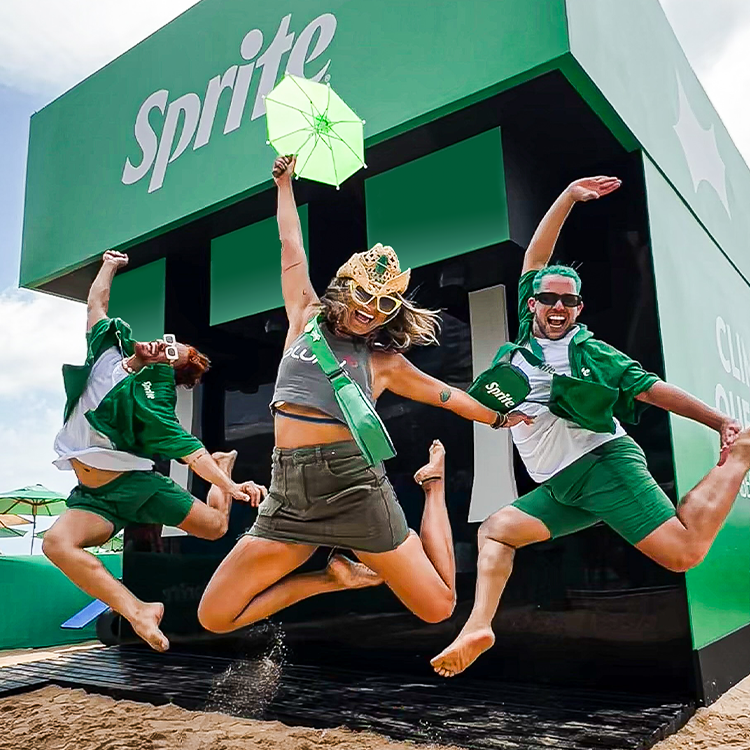 Três pessoas pulam alegremente em frente a duchas gratuitas da Sprite em uma praia. A pessoa no centro usa uma coroa de palha e segura um pequeno guarda-sol verde claro. Todos os três vestem variações de roupas verdes com o tema Sprite.