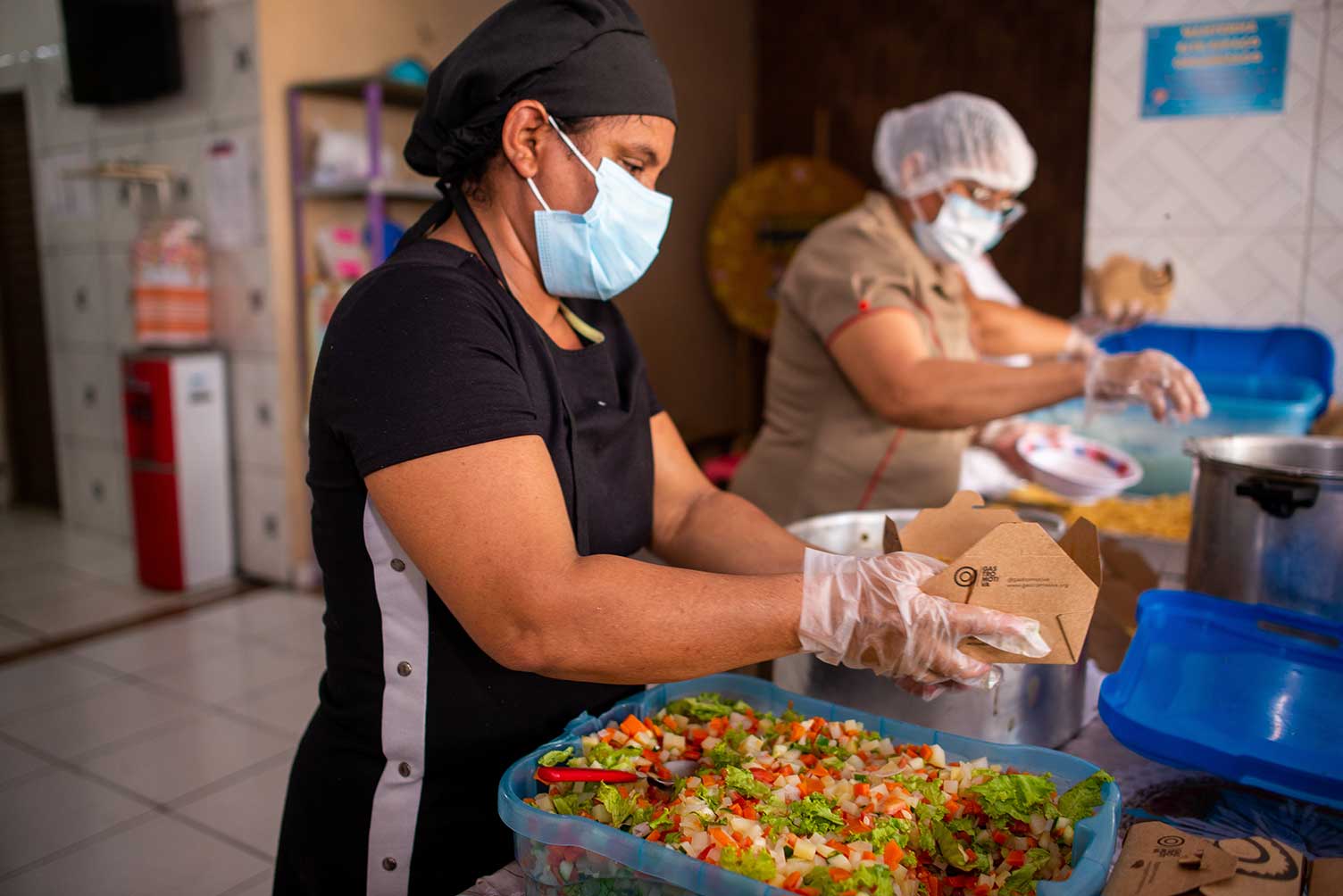 As pessoas que trabalham na cozinha usam máscaras, luvas e toucas de cabelo. Na mesa há comida e panelas.