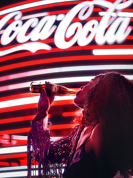 Illuminated Coca-Cola Logo in background, person drinking bottle of Coca-Cola in foreground
