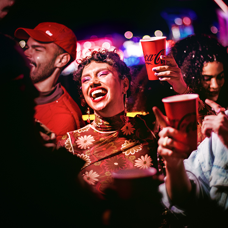 n grupo de amigos de fiesta por la noche, sosteniendo vasos de Coca-Cola y bailando. Una mujer con el pelo rizado, vistiendo una blusa de patrón floral, está en primer plano riéndose alegremente, rodeada de amigos en un entorno animado y colorido.