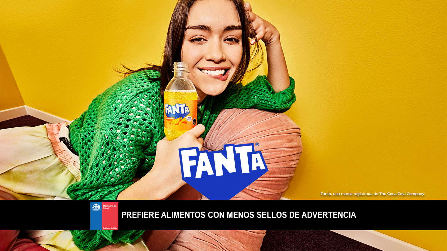 Una joven sonríe con una botella de Fanta Naranja, vestida con un suéter verde y recostada sobre un almohadón en una habitación amarilla.