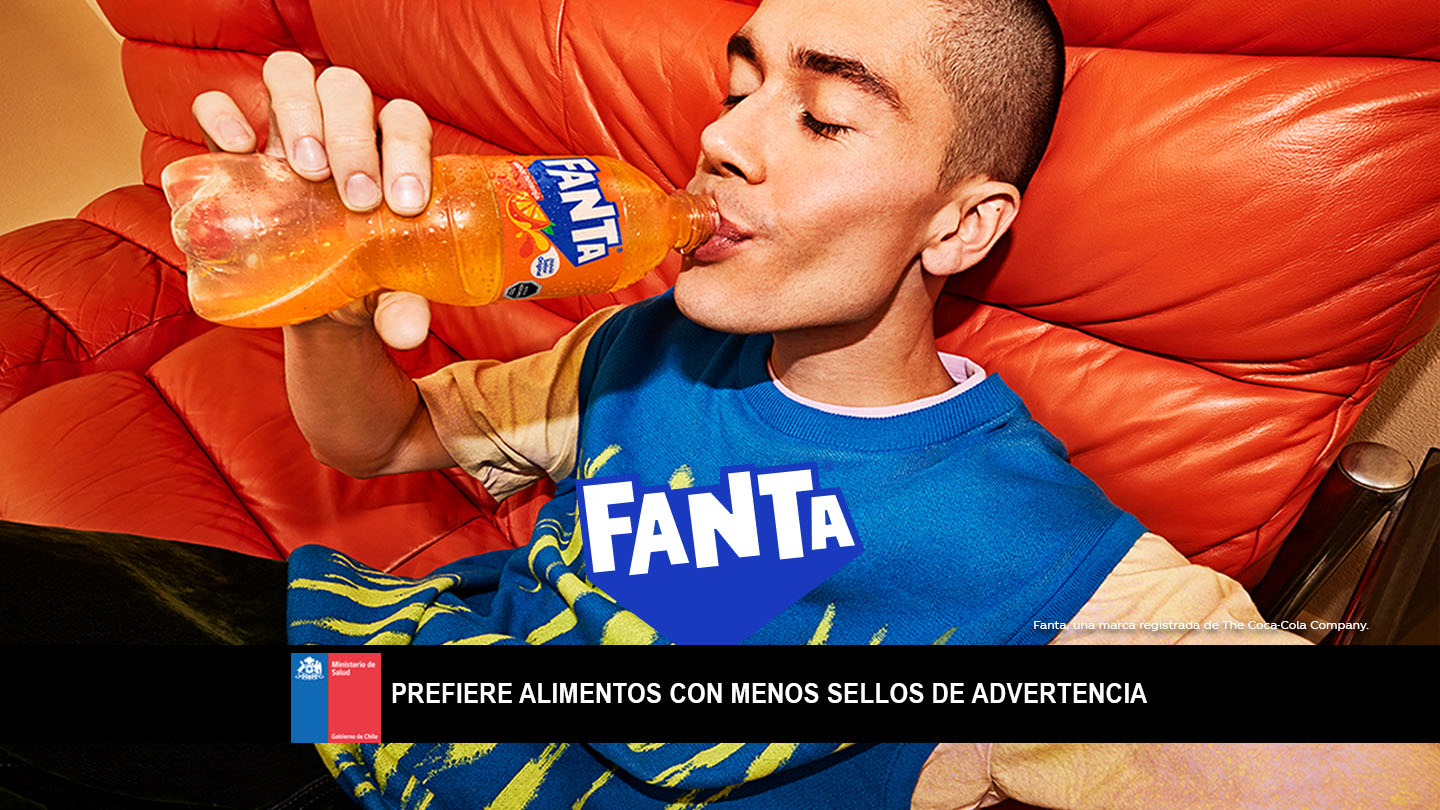 Un joven con una polera azul bebe una botella de Fanta Naranja  recostado en un sillón rojo.