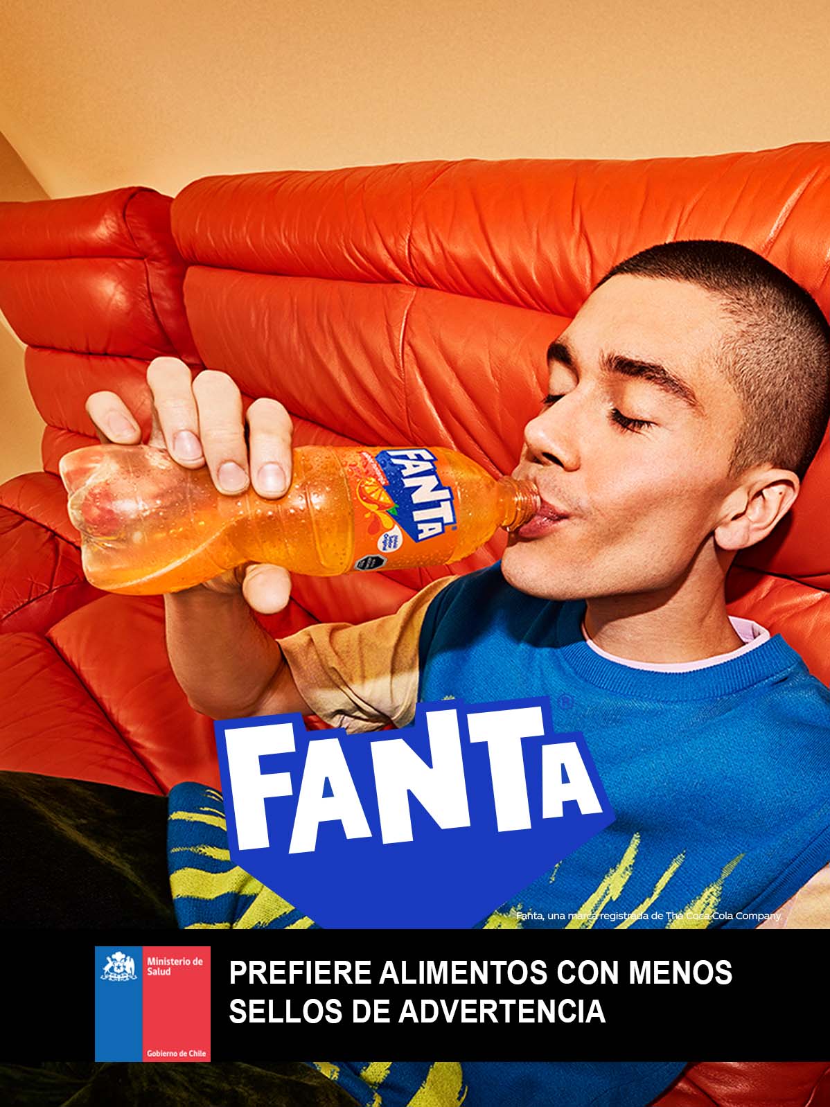 Un joven con una polera azul bebe una botella de Fanta Naranja  recostado en un sillón rojo.