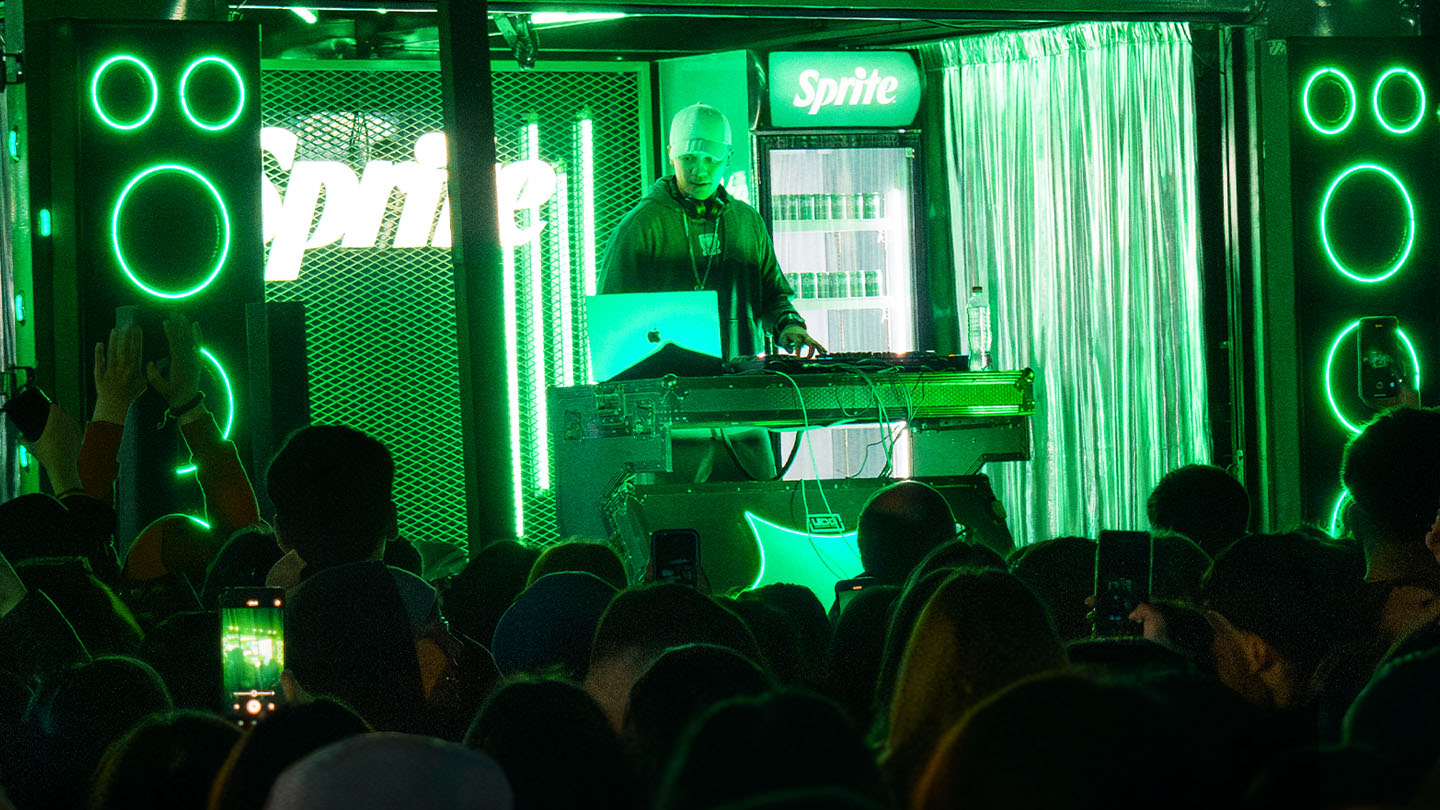 DJ poniendo música sobre el escenario de Sprite RefresCAR con luces verdes, el logo y heladeras de Sprite frente al público.