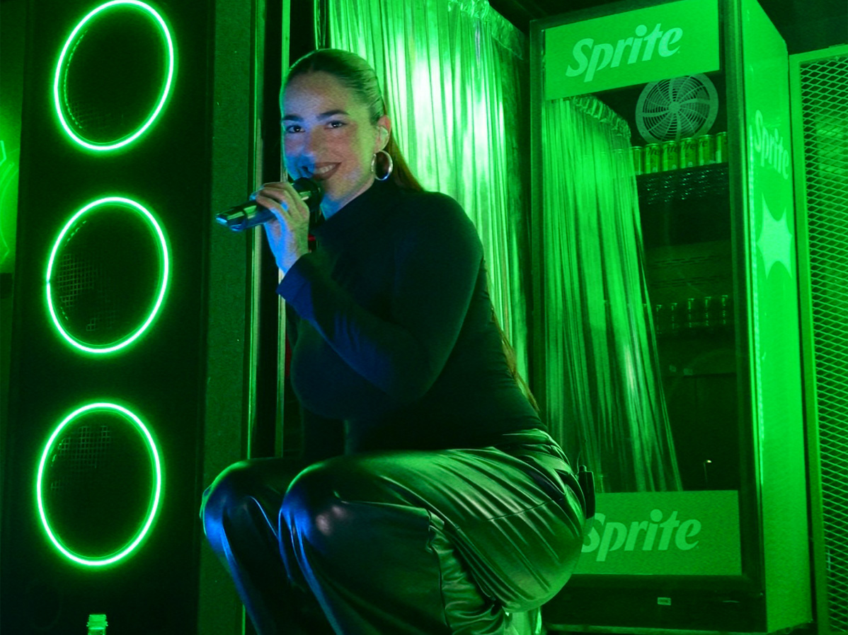 Loyaltty cantando agachada sobre el escenario de Sprite RefresCAR con un fondo de luces verdes y una heladera llena de latas de Sprite frías.