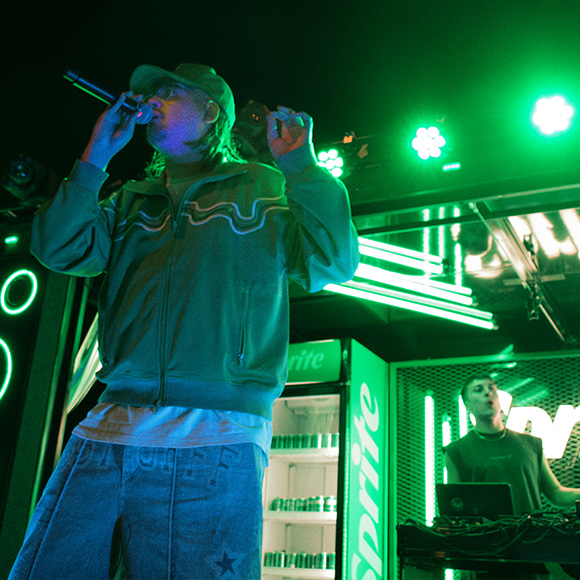 Hombre con gorra cantando en el escenario del Sprite RefresCAR con luces verdes y una heladera de Sprite detrás.