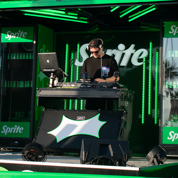 Dj tocando música con su consola sobre el escenario del concierto de Sprite RefresCAR, en un día soleado con las heladeras de Sprite de fondo.