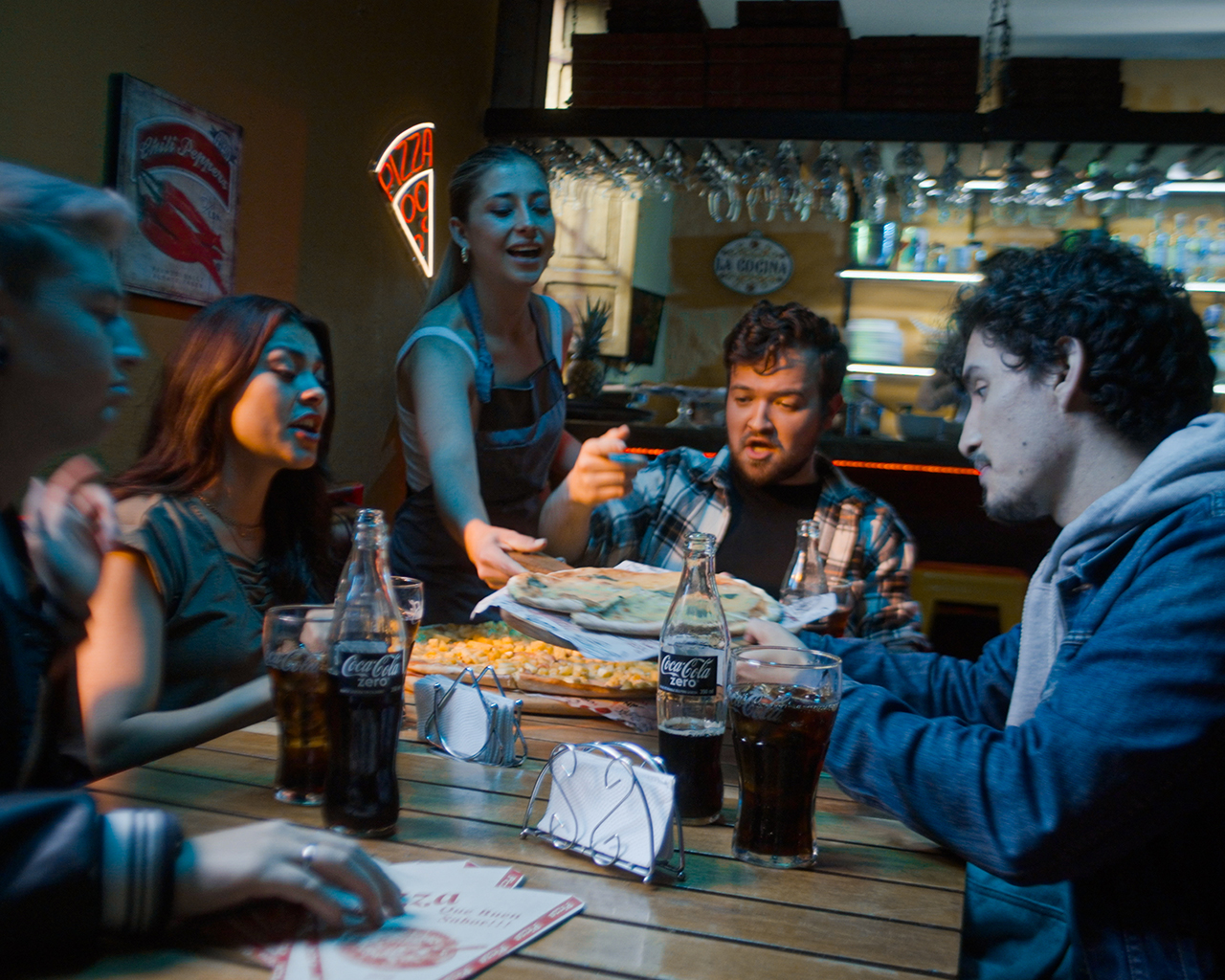 Un grupo de amigos disfrutan de una cena, sobre la mesa hay botellas de Coca-Cola.