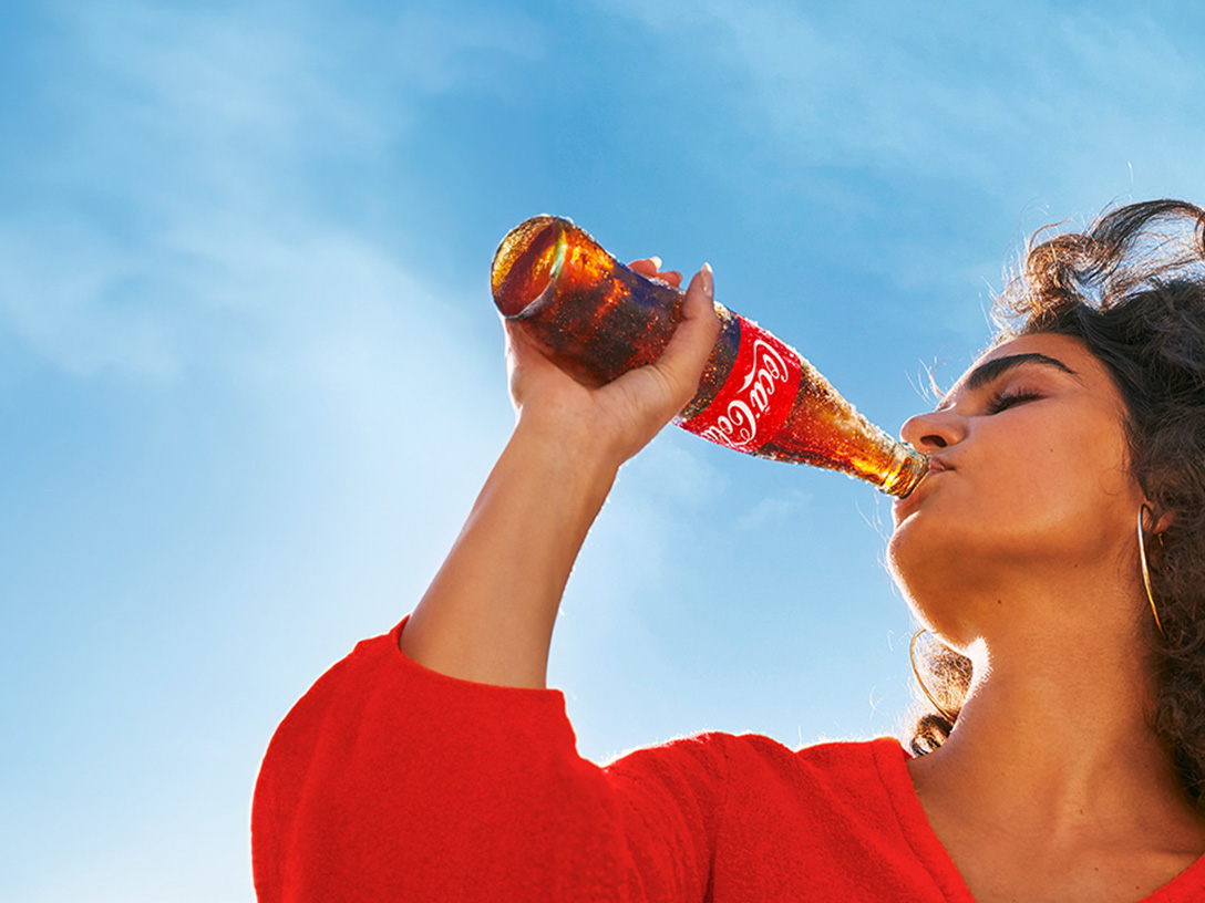 Una mujer con camisa roja bebiendo de una botella de Coca-Cola de vidrio helada en un día soleado.