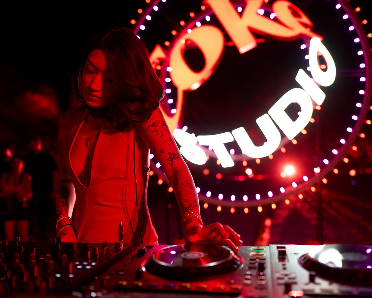 Peggy Gou  tocando en vivo en un evento de Coke Studio, con iluminación vibrante en rojo y el logo al fondo. Lleva un vestido rojo y tiene tatuajes visibles en los brazos. Al fondo, hay personas y un ambiente de fiesta y música.