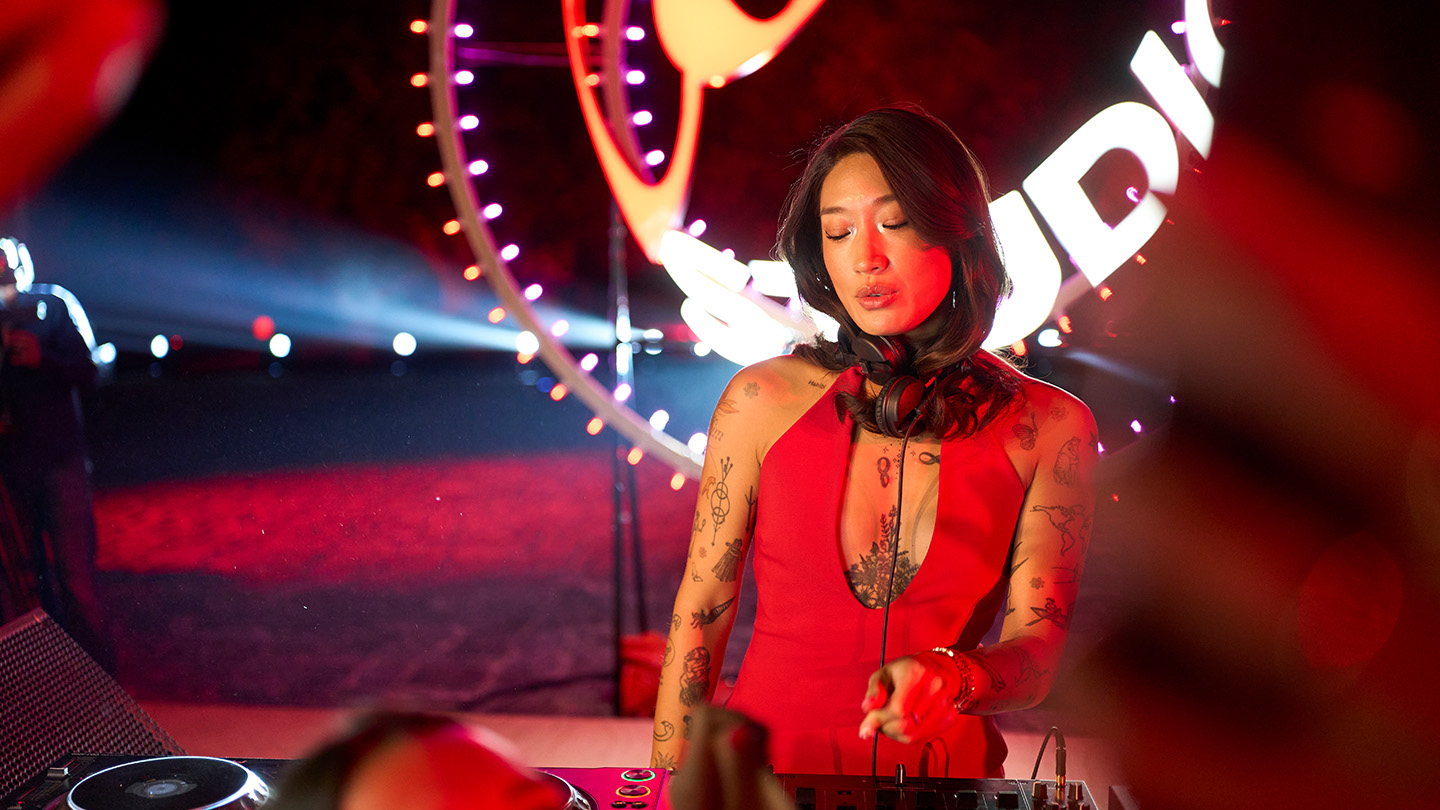 Peggy Gou en un evento de Coke Studio, tocando en un escenario iluminado en rojo, con el logo de fondo. Lleva un vestido rojo y audífonos en el cuello, rodeada de un ambiente de fiesta.