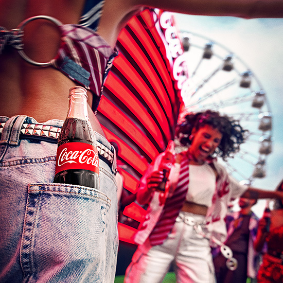  Primer plano de una botella de Coca-Cola en el bolsillo trasero de unos jeans, con una mujer de fondo disfrutando de un festival con una noria y decoraciones en rojo.