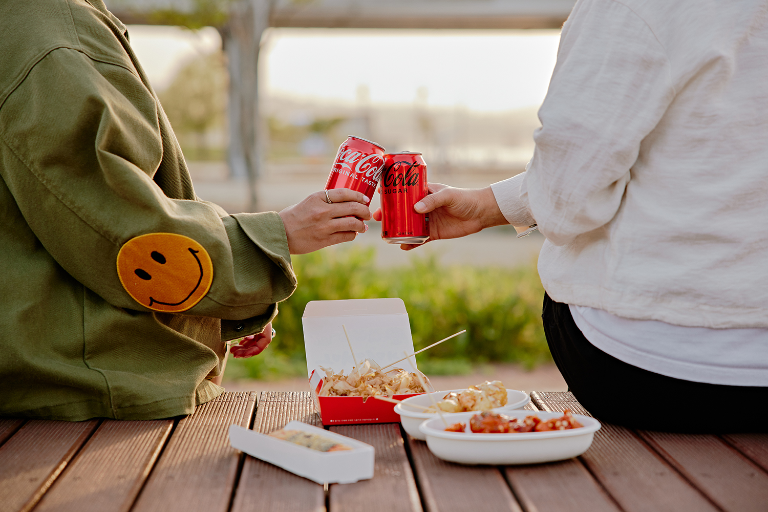데크 위에 야시장 음식을 올려놓고 친구 두 명이서 코카-콜라와 코카-콜라 제로 캔을 건배하는 모습이 담겨있는 이미지이다.