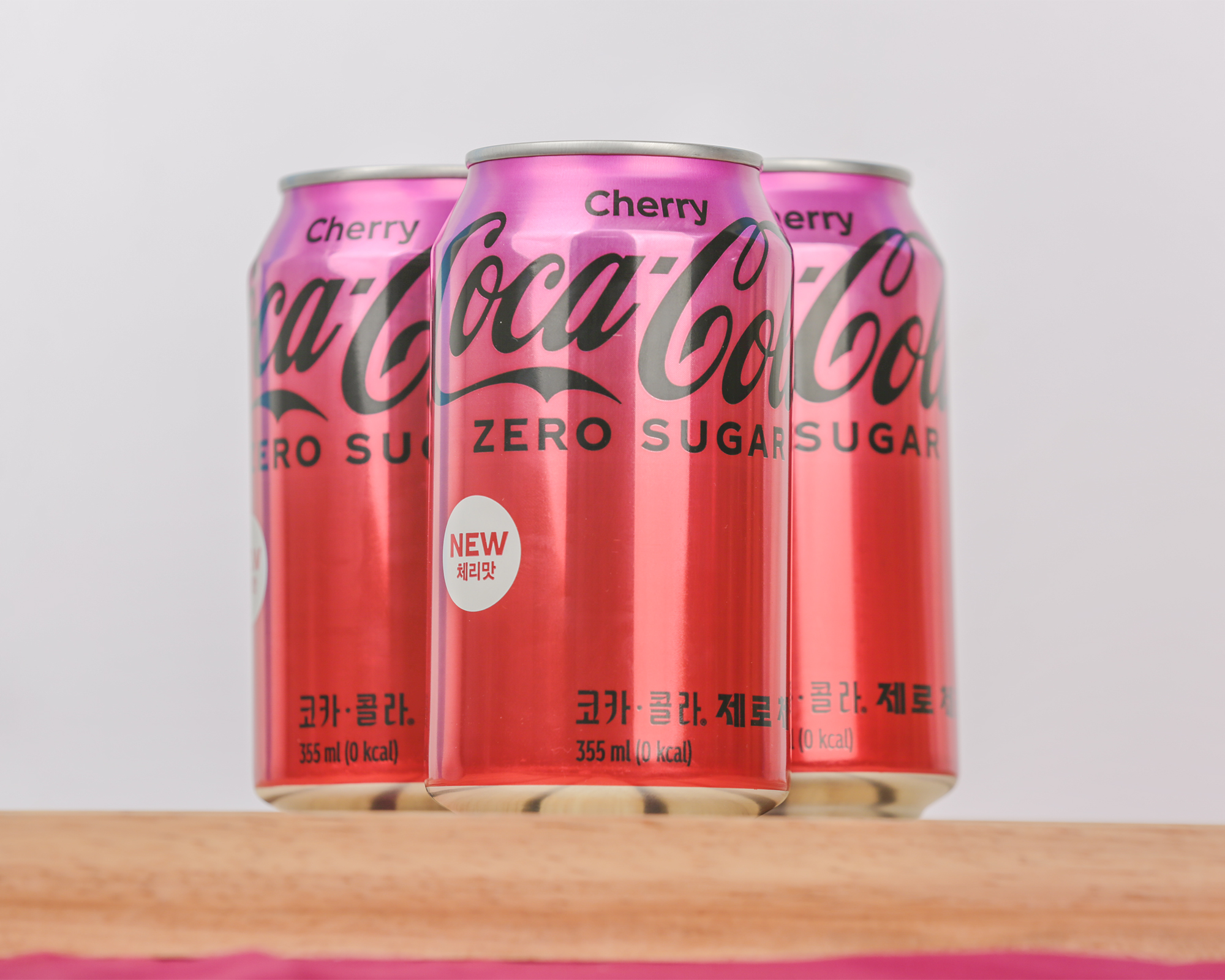 코카-콜라 제로 체리 3캔이 나무 받침대 위에 정렬되어 있다. 제일 앞에 놓여 있는 음료 캔 제품의 좌우 뒷편에 캔이 하나씩 더 놓여져 있다.