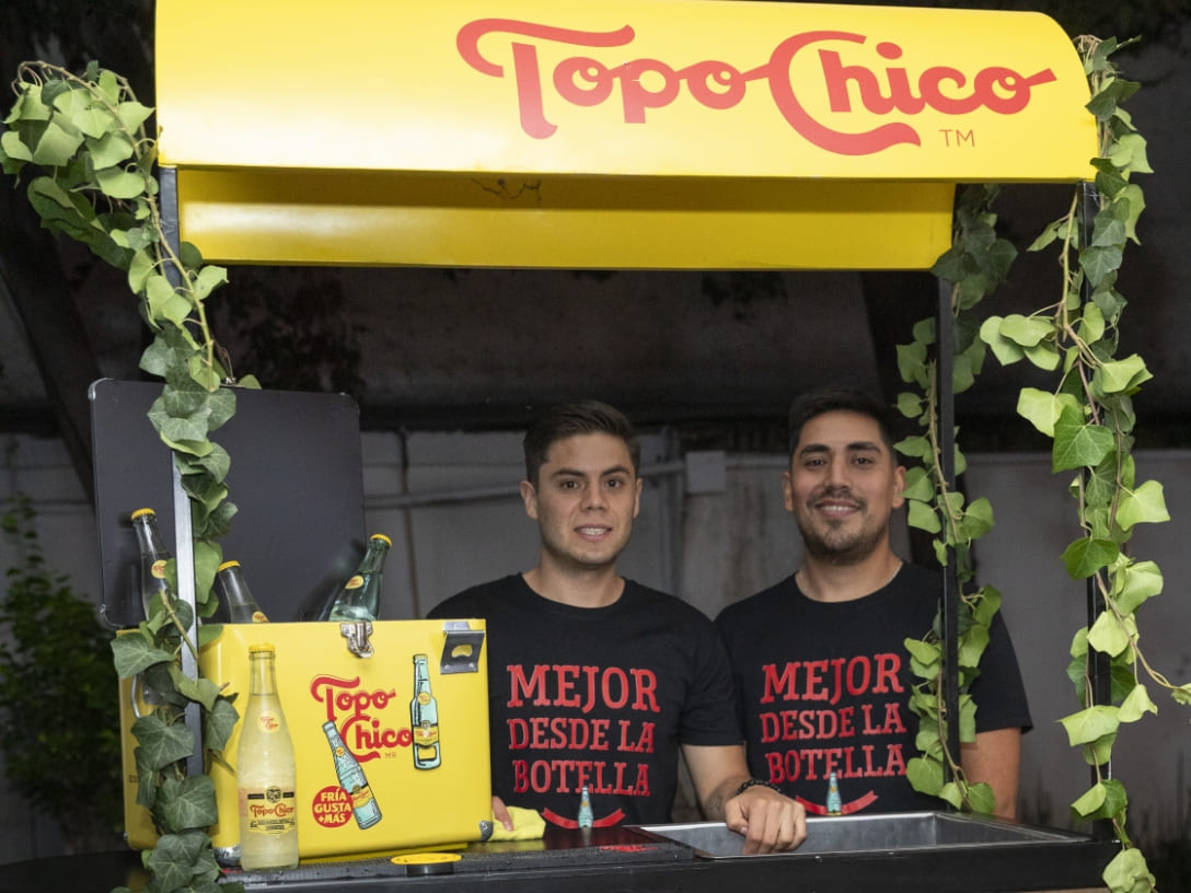 Dos vendedores sonríen detrás de un puesto de Topo Chico, decorado con enredaderas.