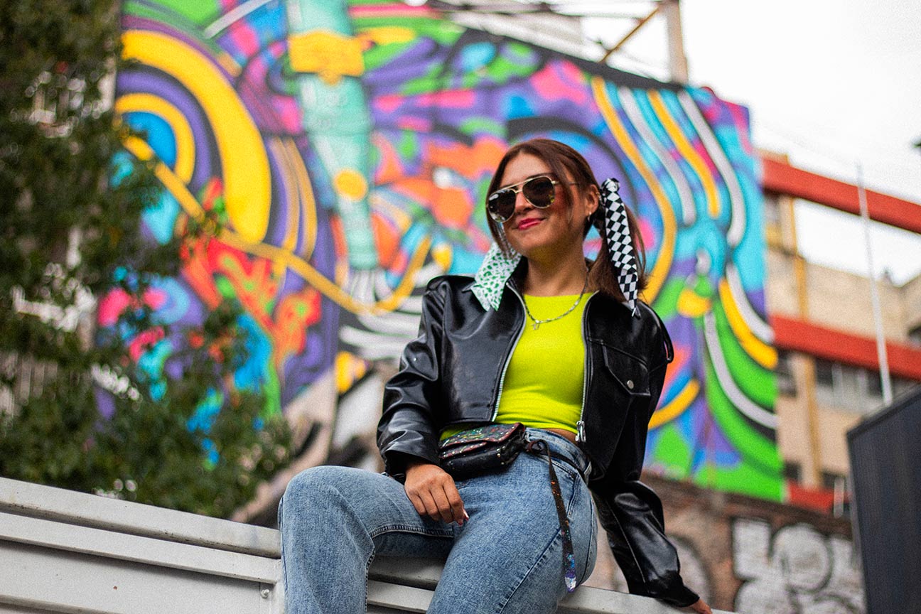 Mujer sentada en frente de un mural de colores. Está utilizando gafas de sol, campera de cuero y remera verde.