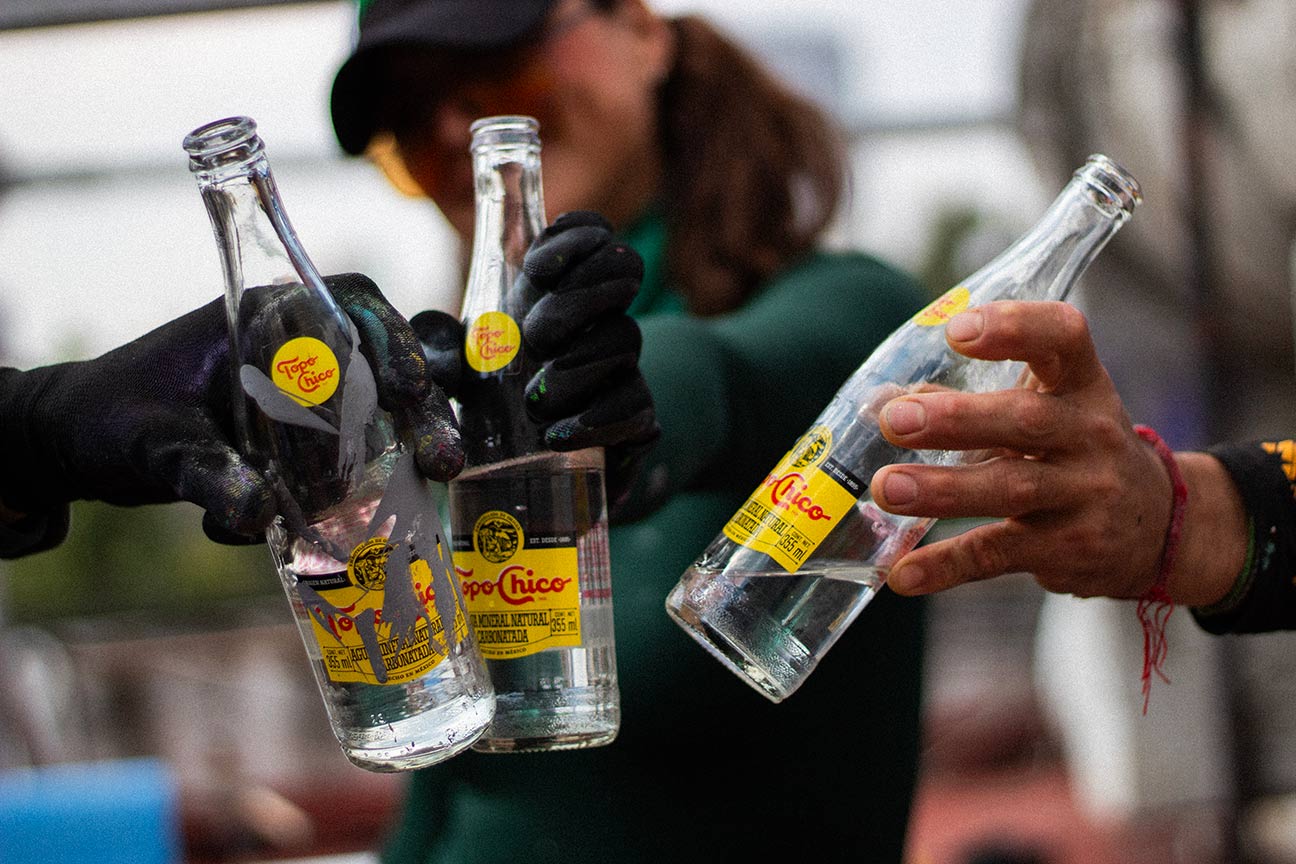 Grupo de tres personas brindando con botellas de vidrio Topo Chico. De fondo se ve desenfocada la cara de una chica.