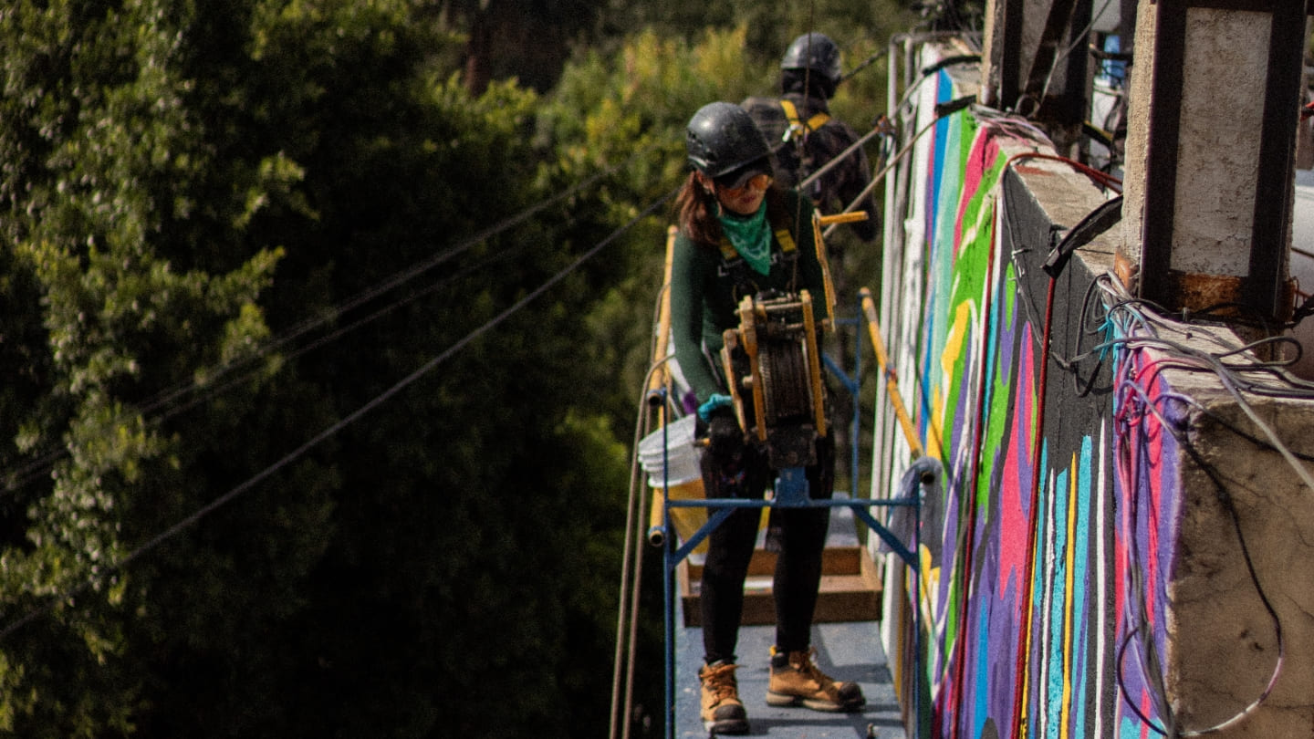 Persona colgada con arnes y casco parada encima de una terraza frente un mural de colores.