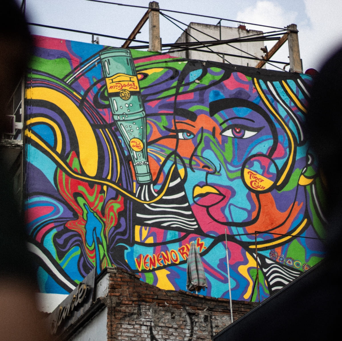 mural de colores con botella Topo Chico. Hay dos personas de espaldas mirando desenfocadas en la imagen