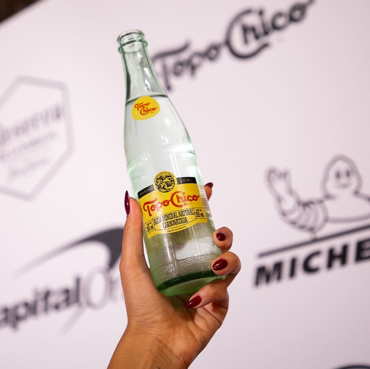 Una mano sostiene una botella de Topo Chico. De fondo, se observa una gráfica con los logos de Topo Chico y Michelin.