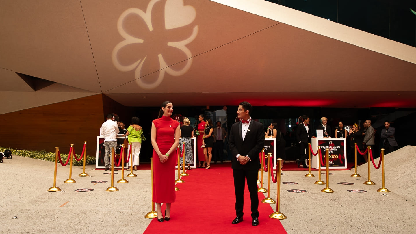 Una mujer con vestido rojo y un hombre de traje reciben a los invitados sobre una alfombra roja. 