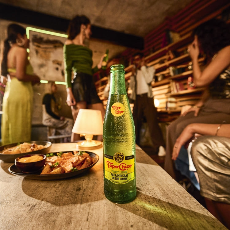 Topo Chico y bocadillos están en una mesa en primer plano, con gente de fiesta y un músico al fondo.