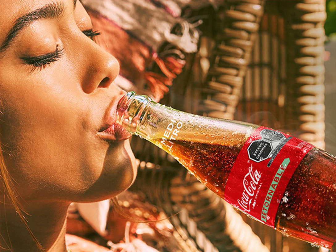 Una mujer bebiendo de una botella de Coca-Cola de vidrio helado en un día soleado, mientras el fondo muestra un tejido de mimbre y algo de vegetación.