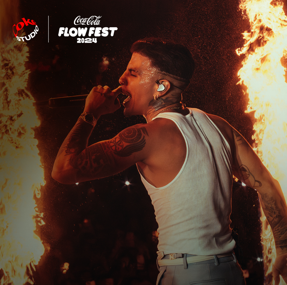 Rauw Alejandro en el escenario del Coca-Cola Flow Fest 2024, sosteniendo un micrófono y cantando apasionadamente mientras está rodeado de columnas de fuego que iluminan el fondo oscuro, transmitiendo energía y emoción.