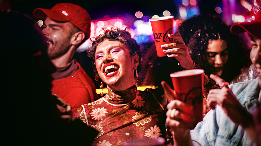 : Un grupo de amigos de fiesta por la noche, sosteniendo vasos de Coca-Cola y bailando. Una mujer con el pelo rizado, vistiendo una blusa de patrón floral, está en primer plano riéndose alegremente, rodeada de amigos en un entorno animado y colorido.