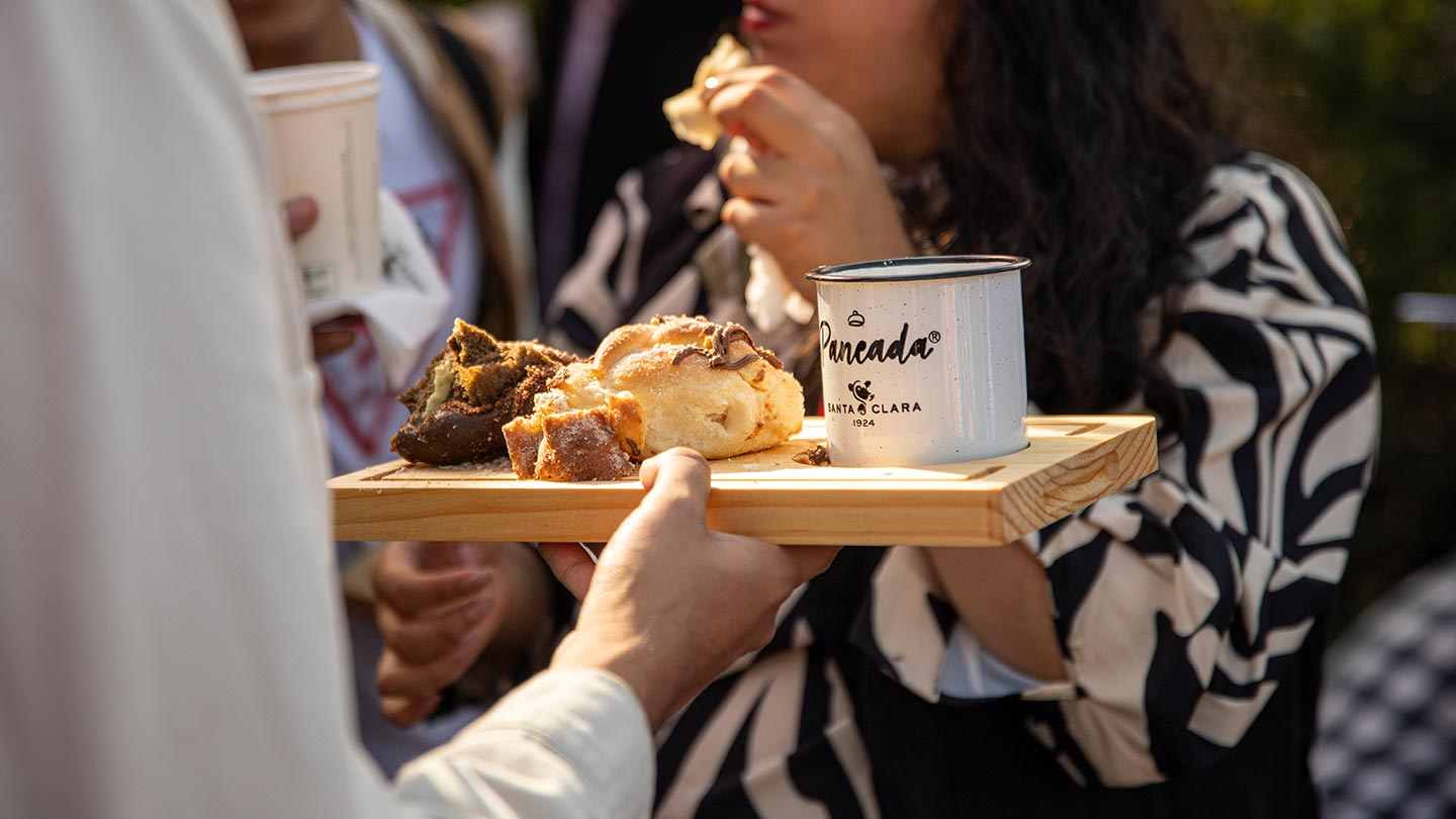 imagen de una persona sosteniendo una tabla con una taza con el texto: Paneada, Santa Clara. En la tabla también hay diferentes preparaciones dulces y de fondo se encuentra una chica comiendo.