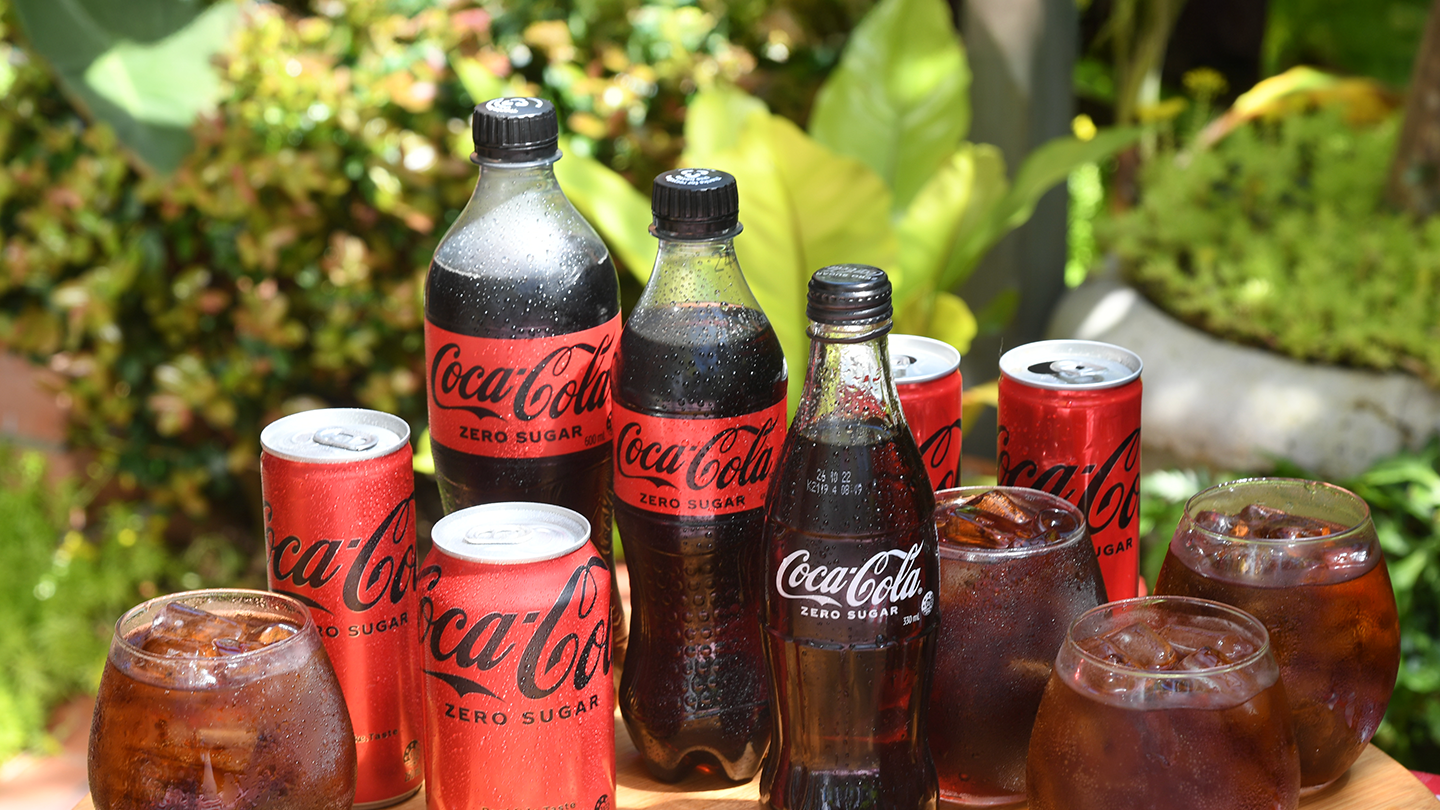 A bottle of Coca-Cola No Sugar, Powerade Zero, and Sprite No Sugar side by side in front of a garden