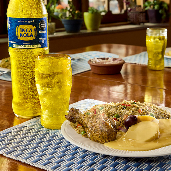 Una botella de Inca Kola Retornable sobre una mesa con 3 platos de comida y dos vasos de vidrio de Inca Kola.