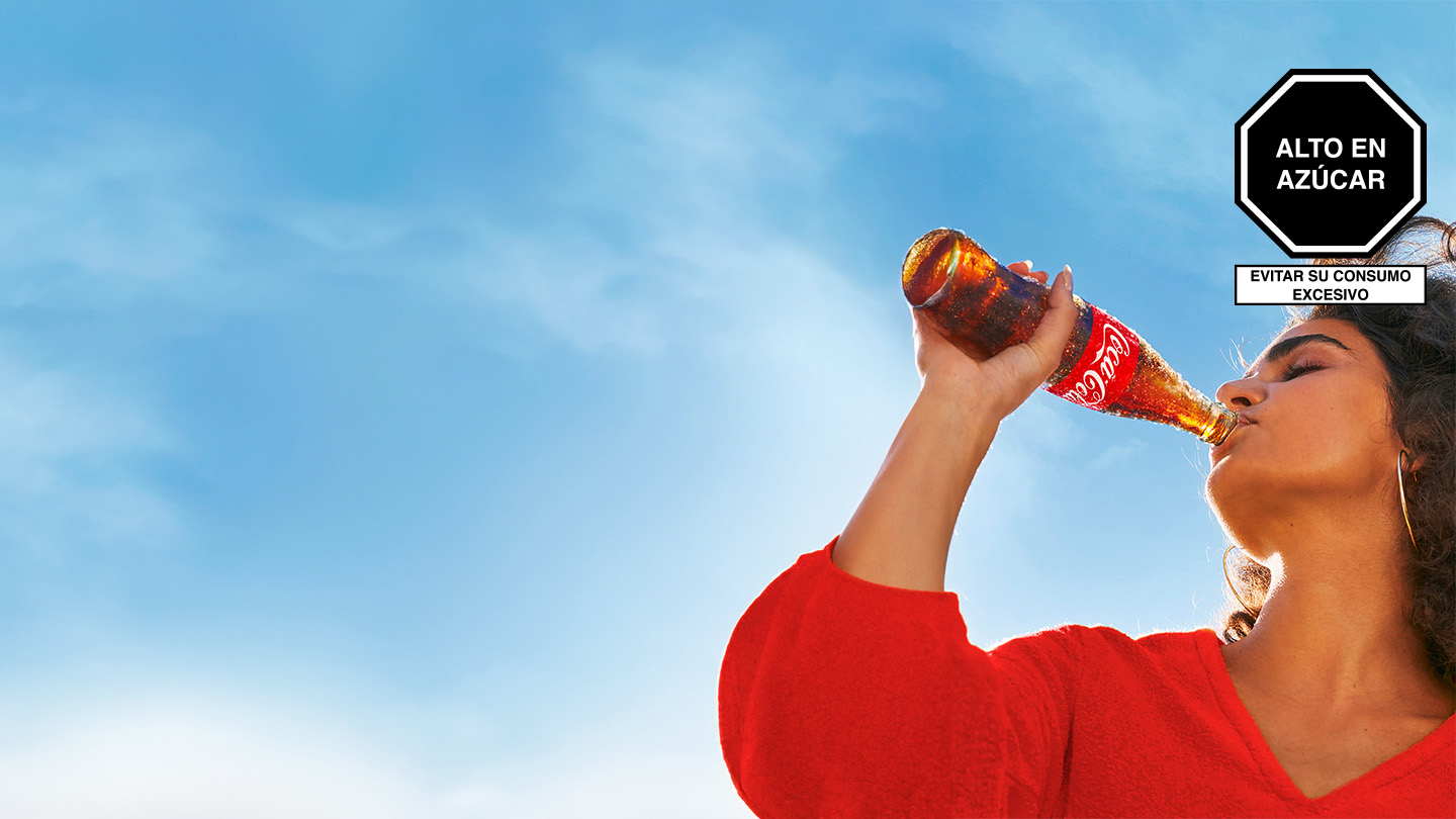 Una mujer con camiseta roja disfruta de una Coca-Cola en botella de vidrio bajo un cielo celeste.