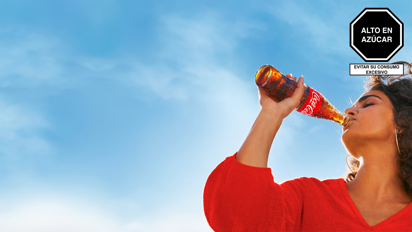 Una mujer con camisa roja bebiendo de una botella de Coca-Cola de vidrio helada en un día soleado.