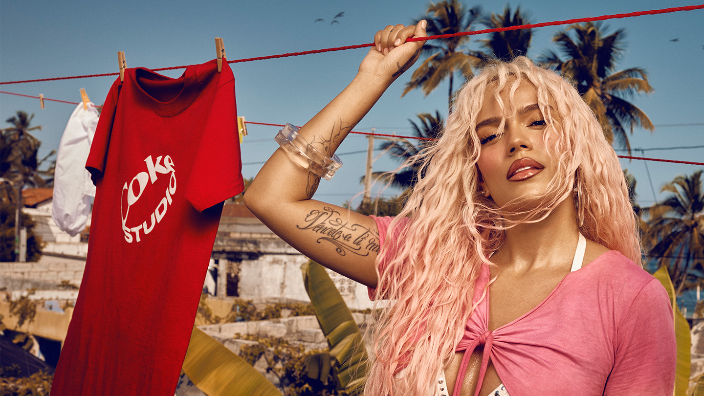 Karol G con el pelo largo y rubio de pie al aire libre cerca de un tendedero con una camiseta roja de Coke Studio colgada. Lleva una parte superior rosa sobre un bikini blanco, mostrando un tatuaje en su brazo. El fondo presenta palmeras y un cielo azul claro.
