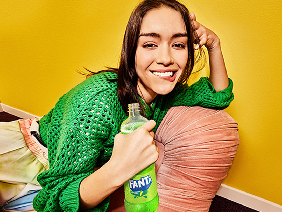 Una joven sonríe con una botella de Fanta Manzana, vestida con un suéter verde y recostada sobre un almohadón en una habitación amarilla.