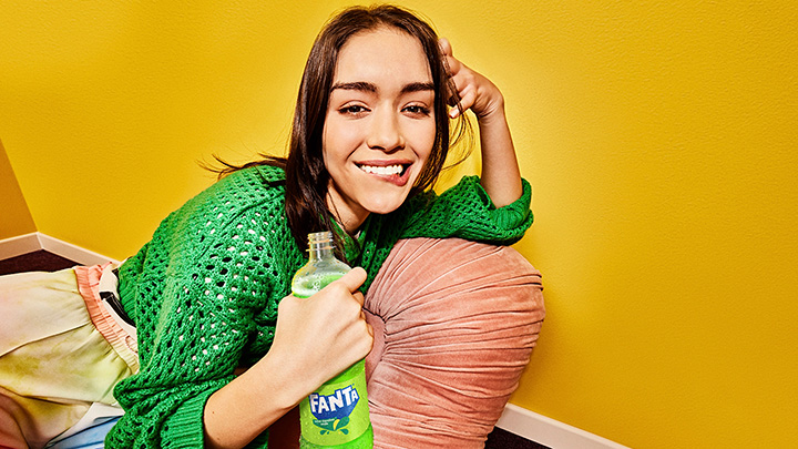 Una joven sonríe con una botella de Fanta Manzana, vestida con un suéter verde y recostada sobre un almohadón en una habitación amarilla.