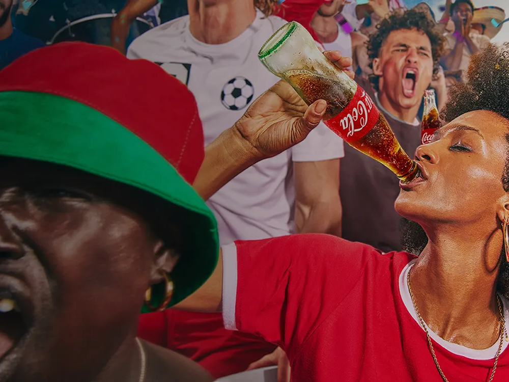 A woman drinks a Coca-Cola from a glass bottle while cheering with other excited soccer fans in the stands during a match.