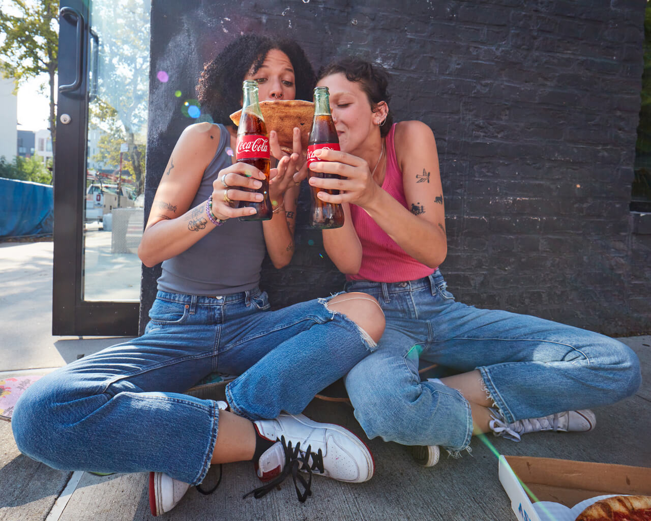 Two women sitting down by a building each holding a Coca-cola