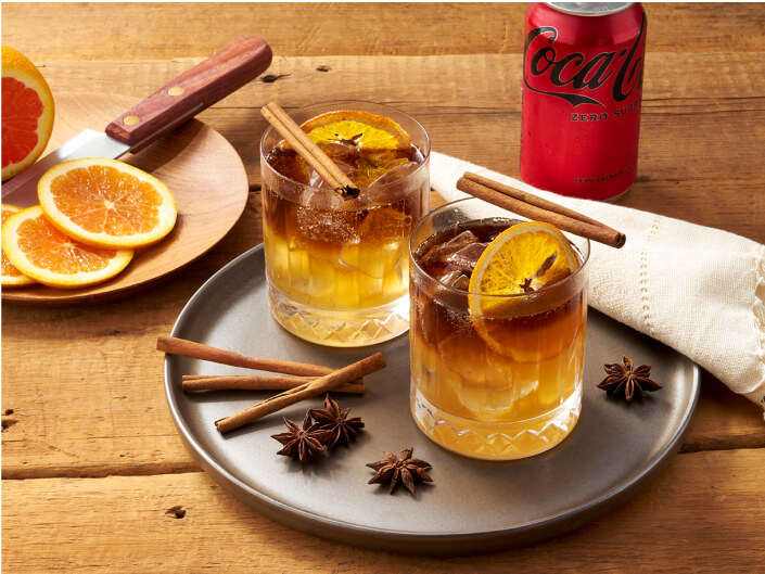 two orange drinks on a plate with cinnamon and cloves beside a coke zero